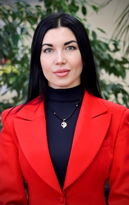 Жінка з довгим чорним волоссям у червоному піджаку стоїть серед рослин.